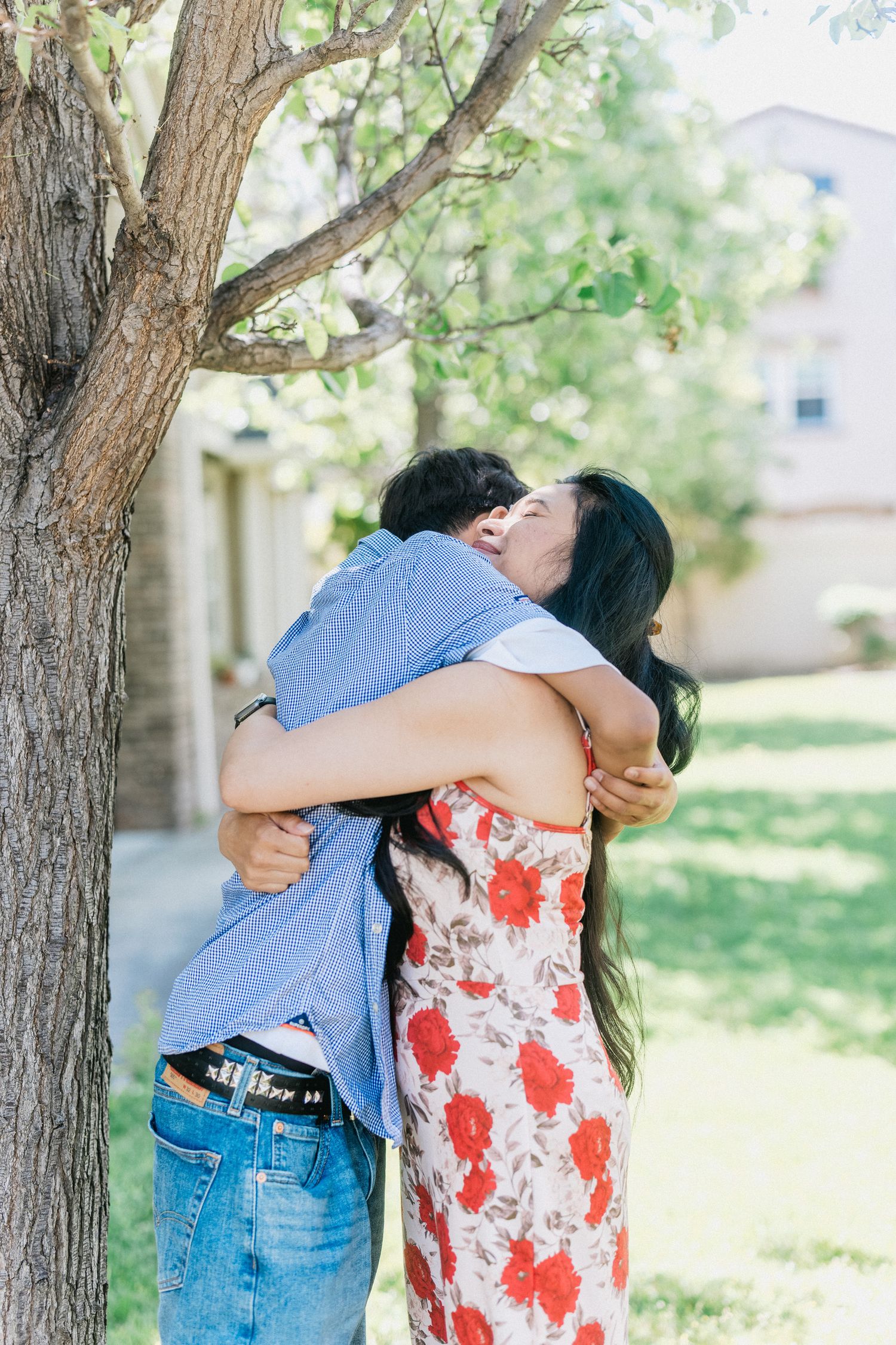 Couple embraces in a tight hug while standing next to a large tree on a sunny day.