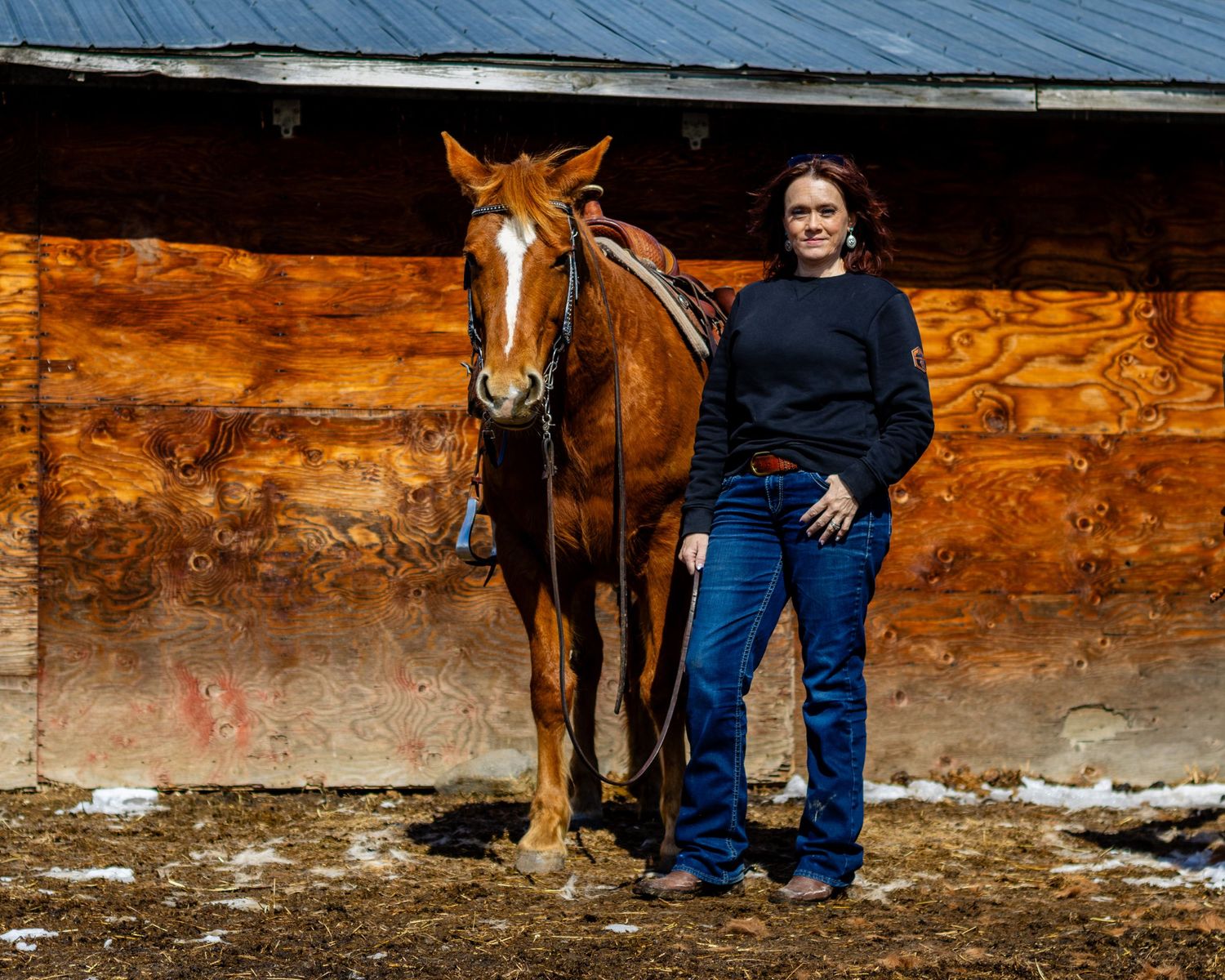 Photographer in black sweater stands with sorrel horse against wooden barn wall.