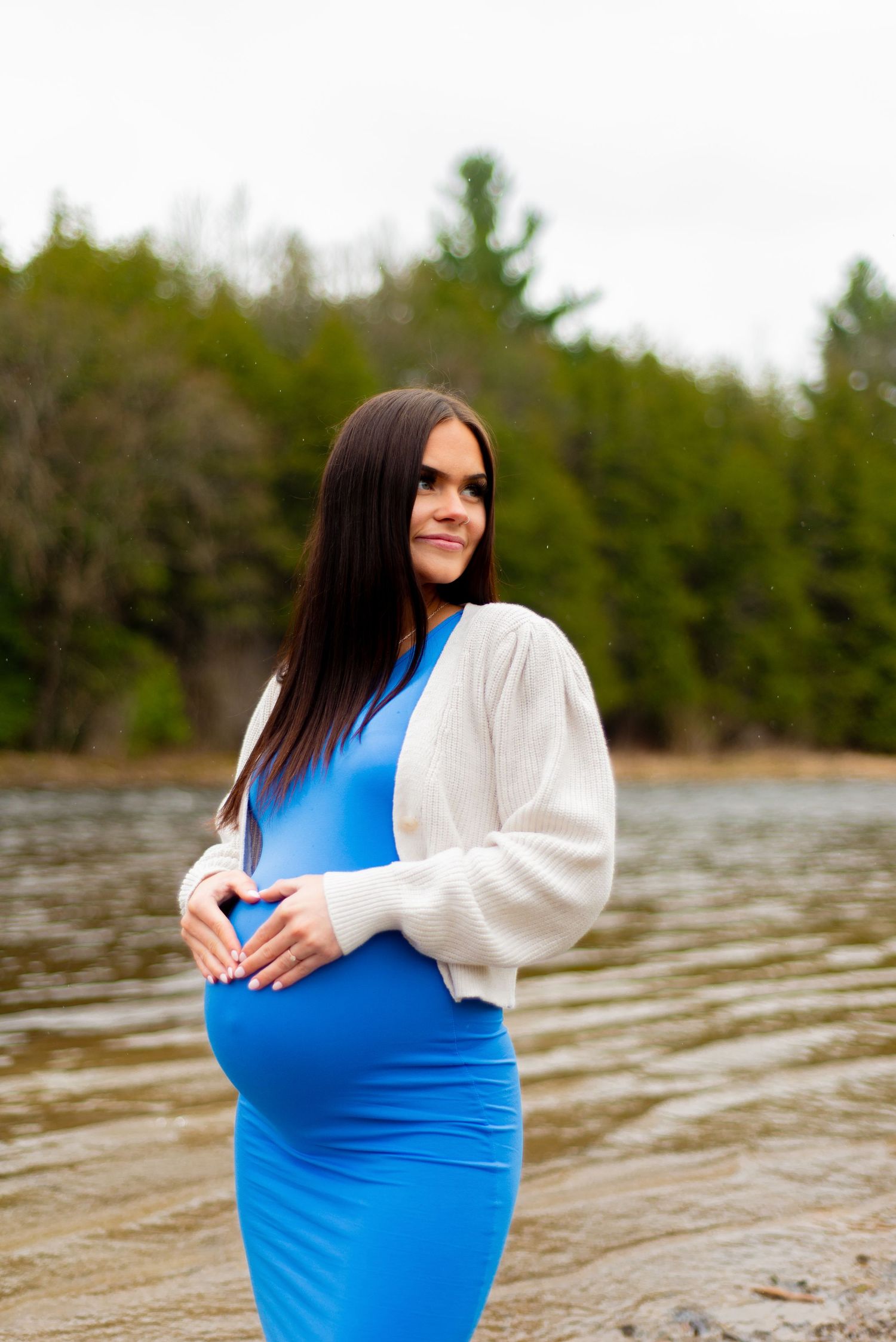 A scenic maternity portrait by a lake with evergreen trees in the background.