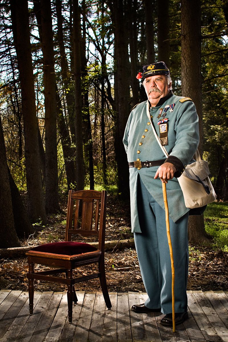 Man in Civil War Confederate officer uniform with cane and medals poses beside a chair in a forest.