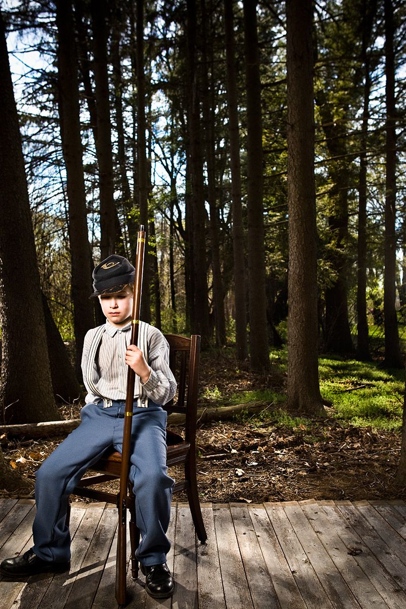 Young boy in Civil War Union soldier uniform sits on a chair holding a rifle in a forest setting.