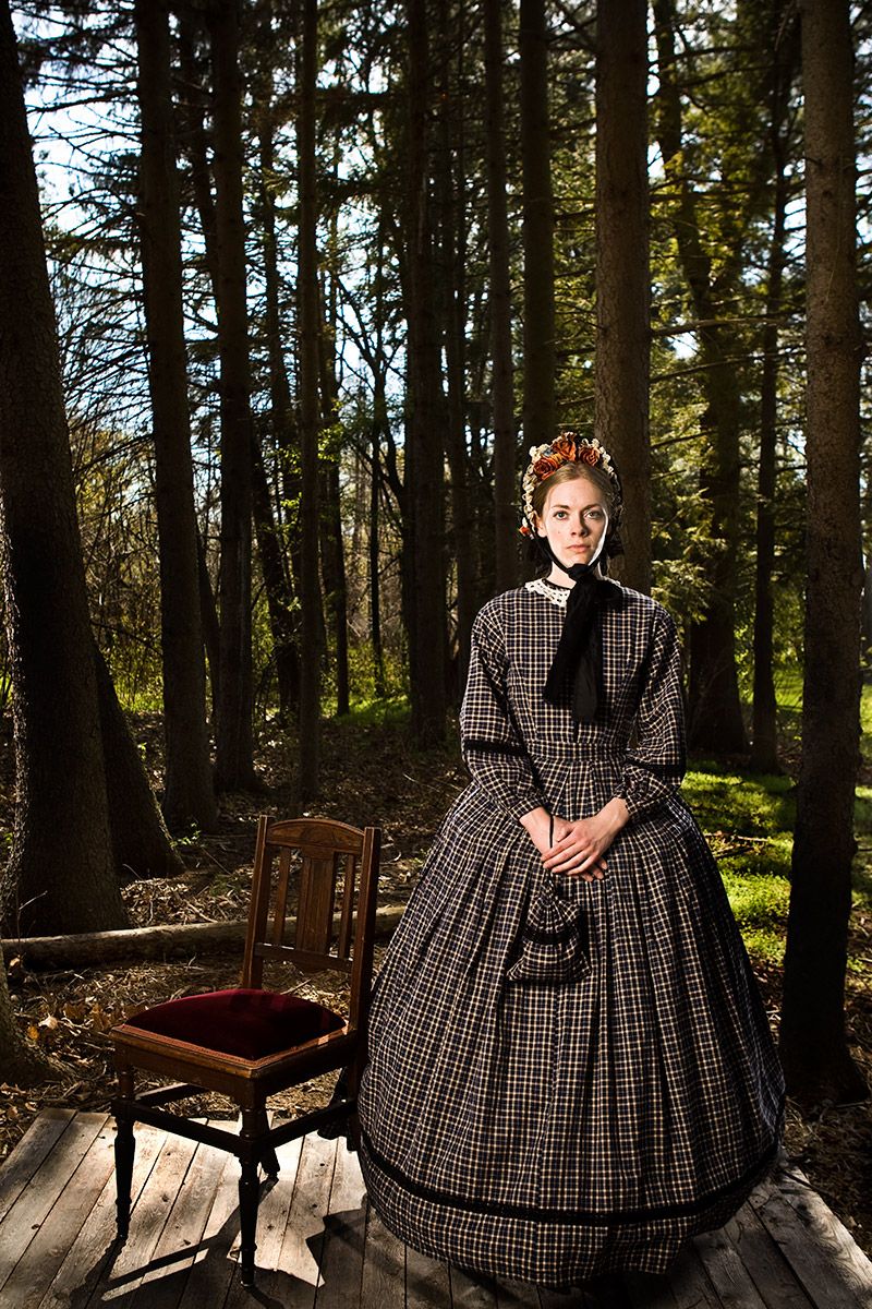 Woman in Victorian plaid dress stands on wooden platform in forest beside antique chair.