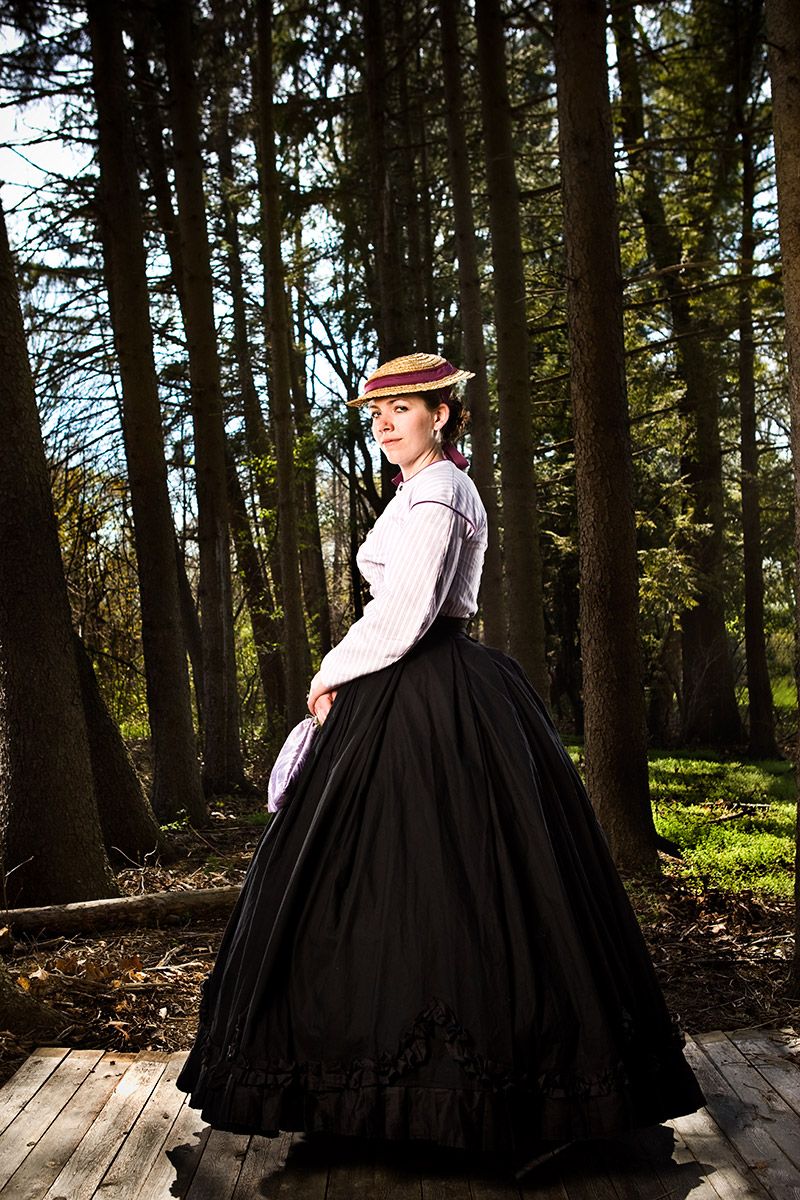 Woman in period costume with straw hat and black skirt poses in sunlit forest setting.