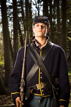 Young Union Civil War soldier in blue uniform holds rifle while standing in forest.