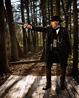 Man in black frontier costume aims pistol on wooden walkway in sunlit forest.