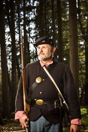 Bearded Union Civil War soldier in dark uniform carries rifle in forest setting.