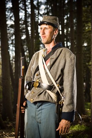 Confederate Civil War soldier in gray uniform stands with rifle in forest setting.