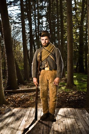 Bearded frontier soldier in worn leather pants holds rifle on wooden platform in woods.