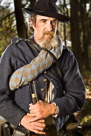Bearded Civil War soldier in gray coat and black hat holds rifle in woods.