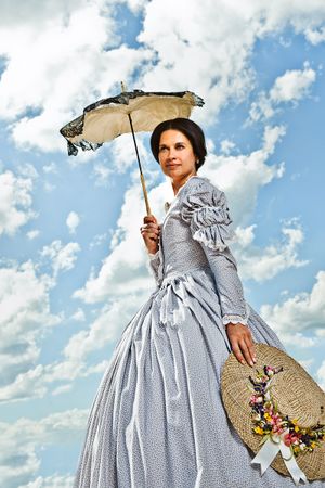 Victorian woman in gray period dress holding parasol and floral hat poses against cloudy blue sky.
