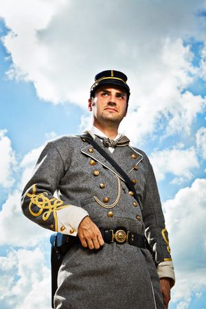 Confederate Civil War officer in gray uniform with gold buttons stands against cloudy blue sky.