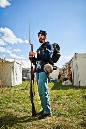 Union Civil War soldier in blue uniform holds rifle with bayonet at Civil War reenactment camp.