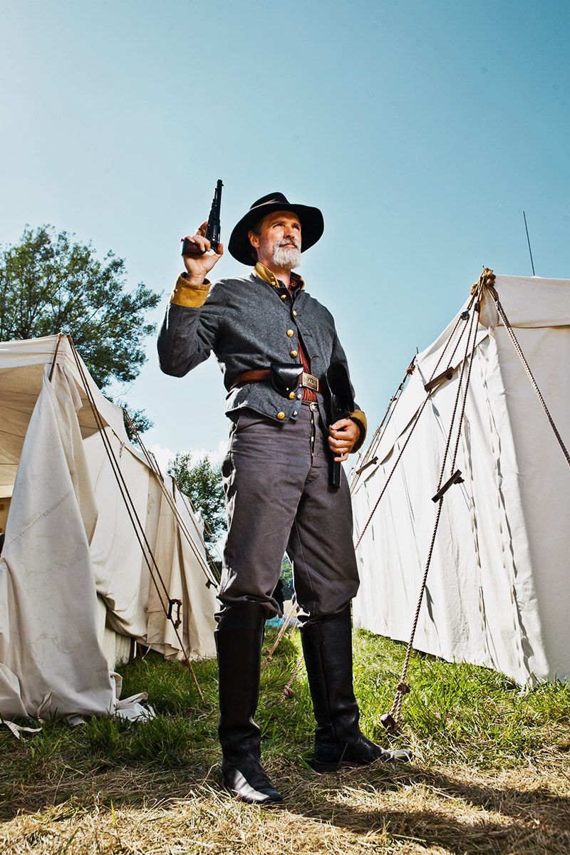 Confederate Civil War reenactor in gray uniform raises pistol between white tents at outdoor camp.