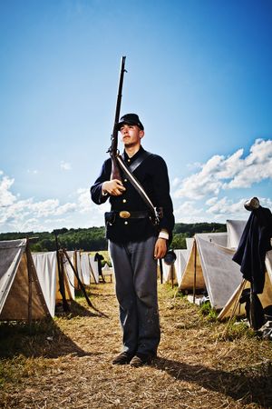 Young Union Civil War soldier holds rifle upright at reenactment camp with white tents.