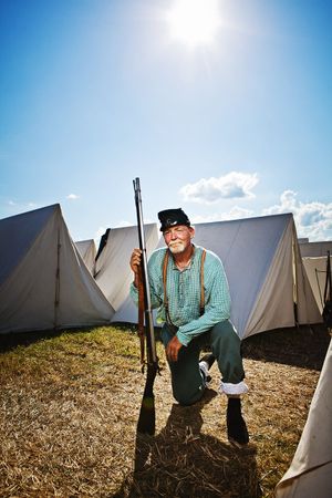 Elderly Civil War reenactor kneels with rifle at sunny outdoor camp with white canvas tents.