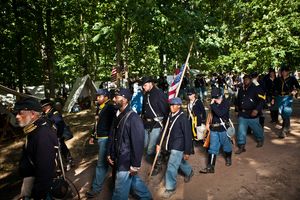 Union Civil War soldiers march in formation carrying flags through wooded reenactment encampment.