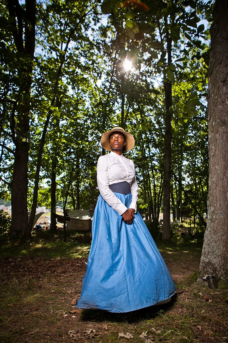 African American woman in period blue skirt and white blouse stands in sunlit forest clearing.