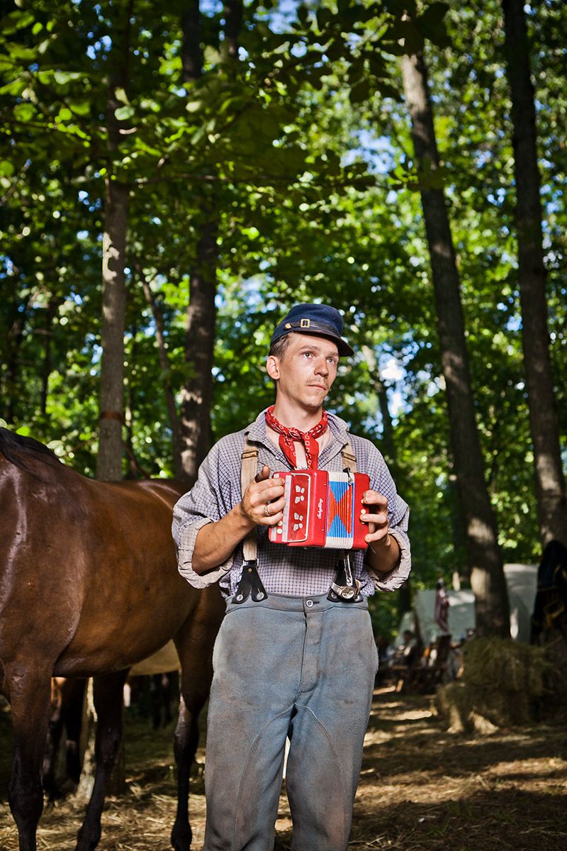 Man in period costume holding a red accordion near a horse in a forest setting.
