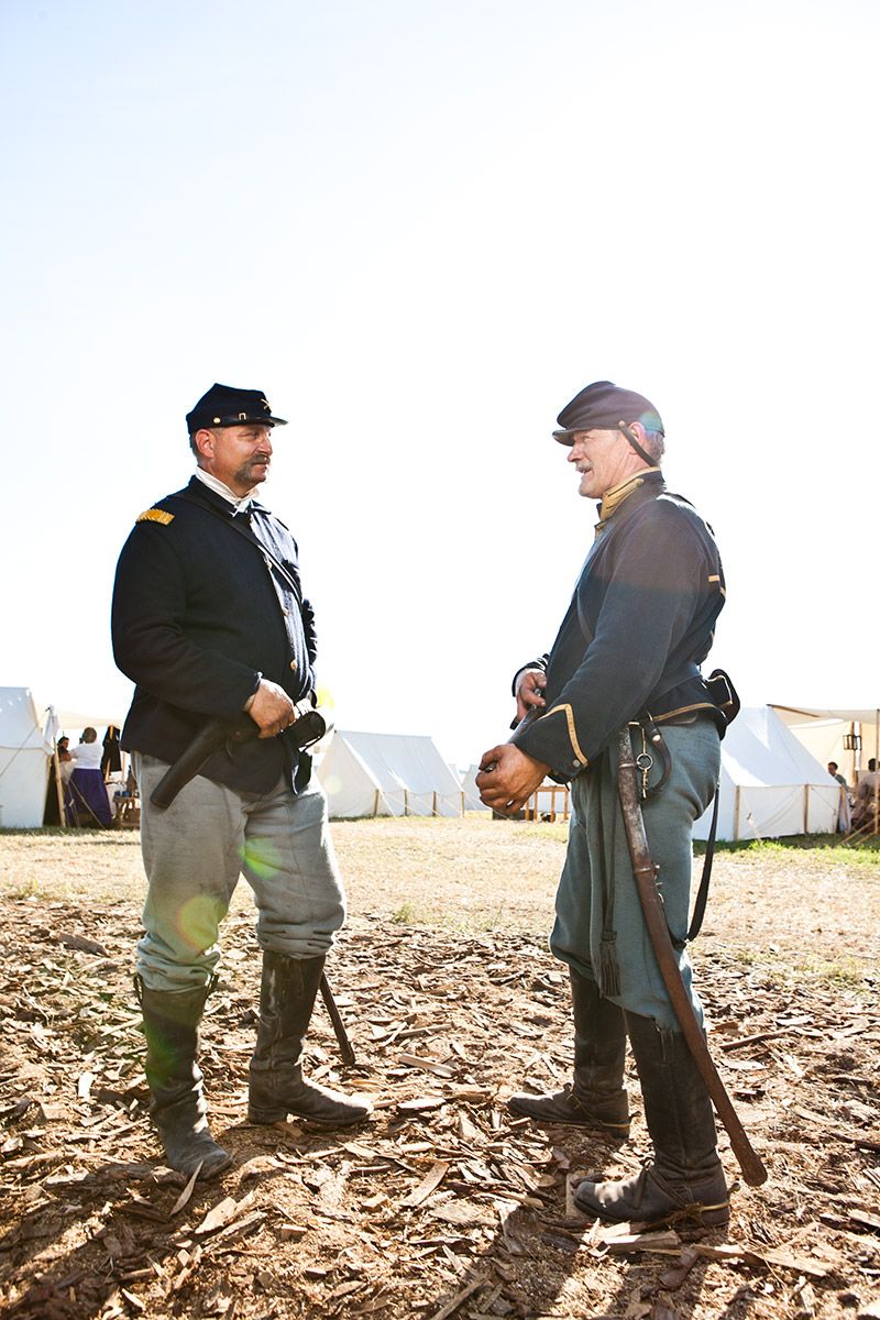 Two Civil War reenactors in Union uniforms converse at outdoor reenactment camp with white tents.