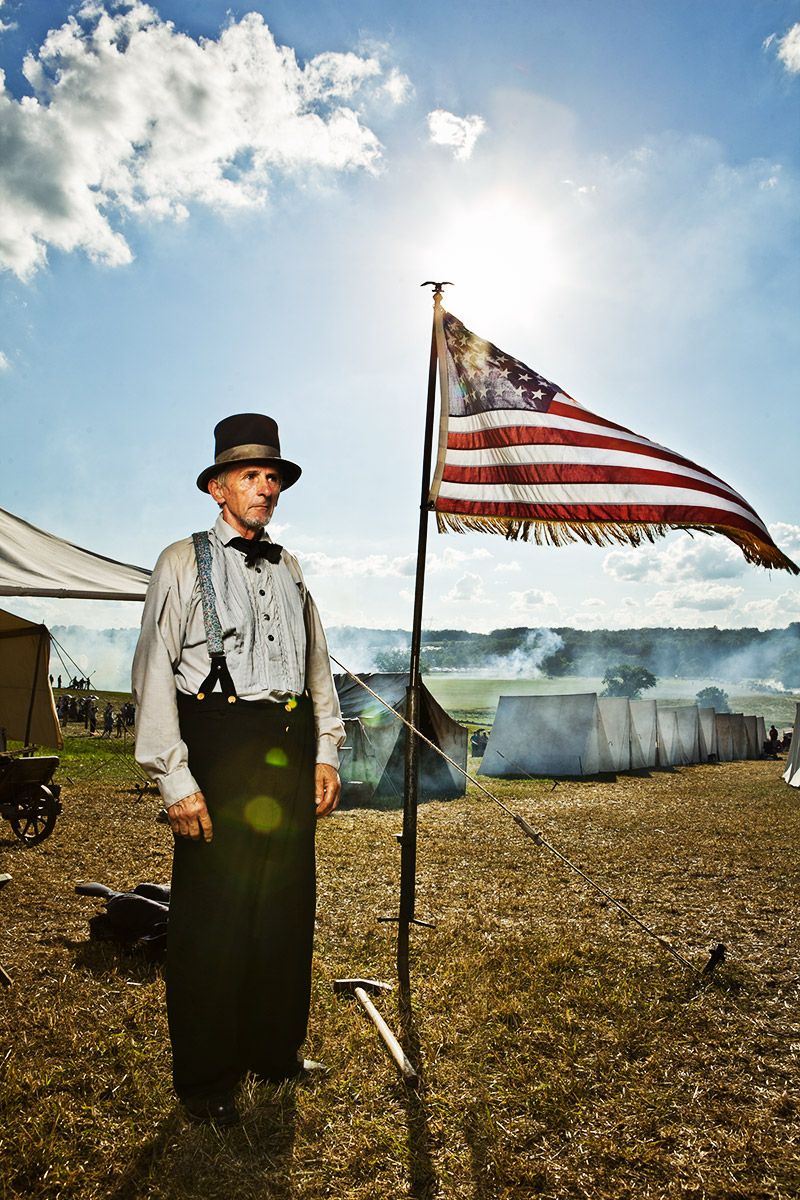 Civil War reenactor in top hat standing beside an American flag at an outdoor event.