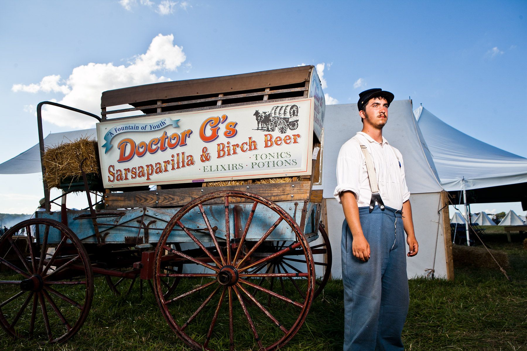 Young man in suspenders standing beside a Doctor G's Sarsaparilla wagon at a fair.
