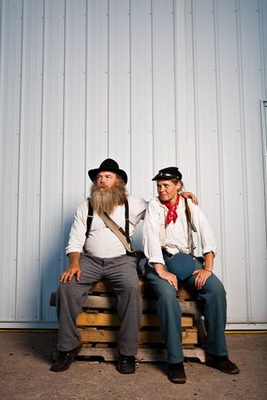 Two people in vintage western costumes sitting on wooden pallets against a metal wall.