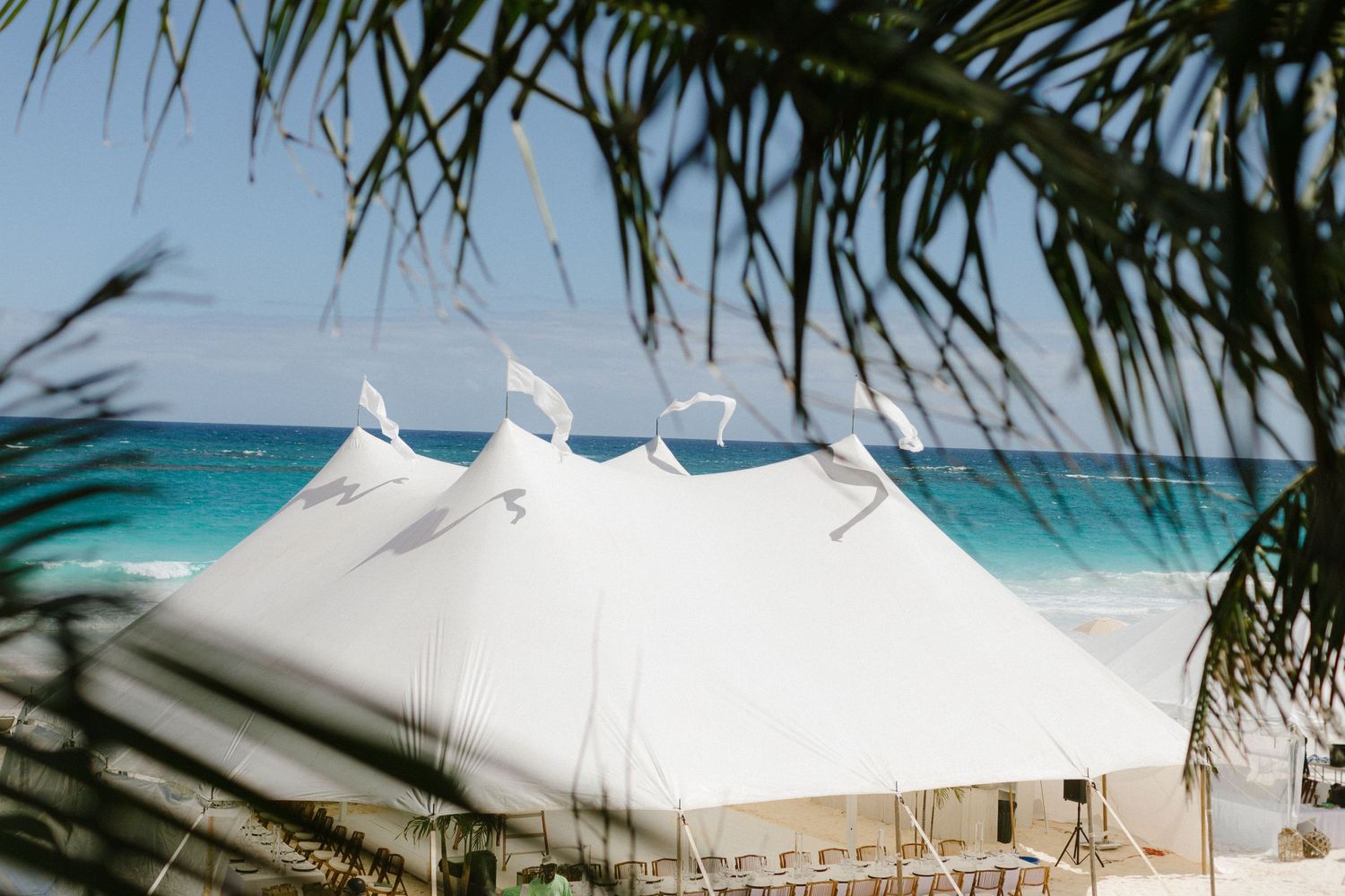 Large white wedding tent set up on a tropical beach with turquoise waters and palm fronds in view.