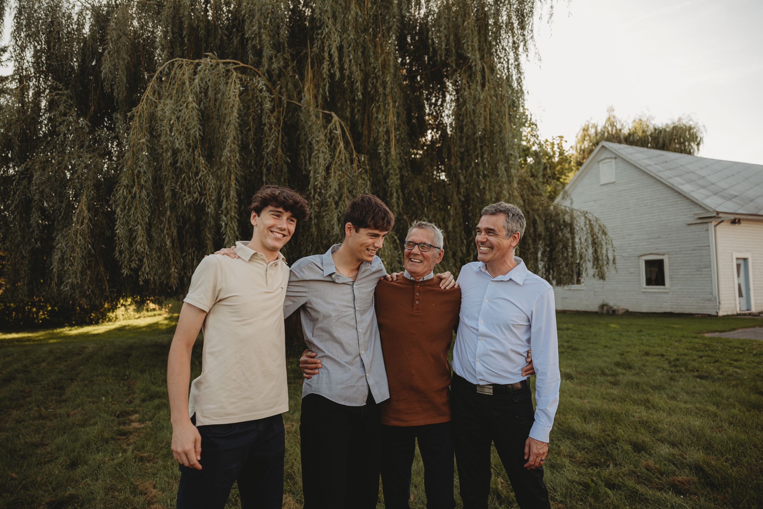 Four friends pose together outdoors with a white building and willow tree in background.