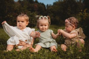 Three babies sitting together in grass wearing light colored outfits.