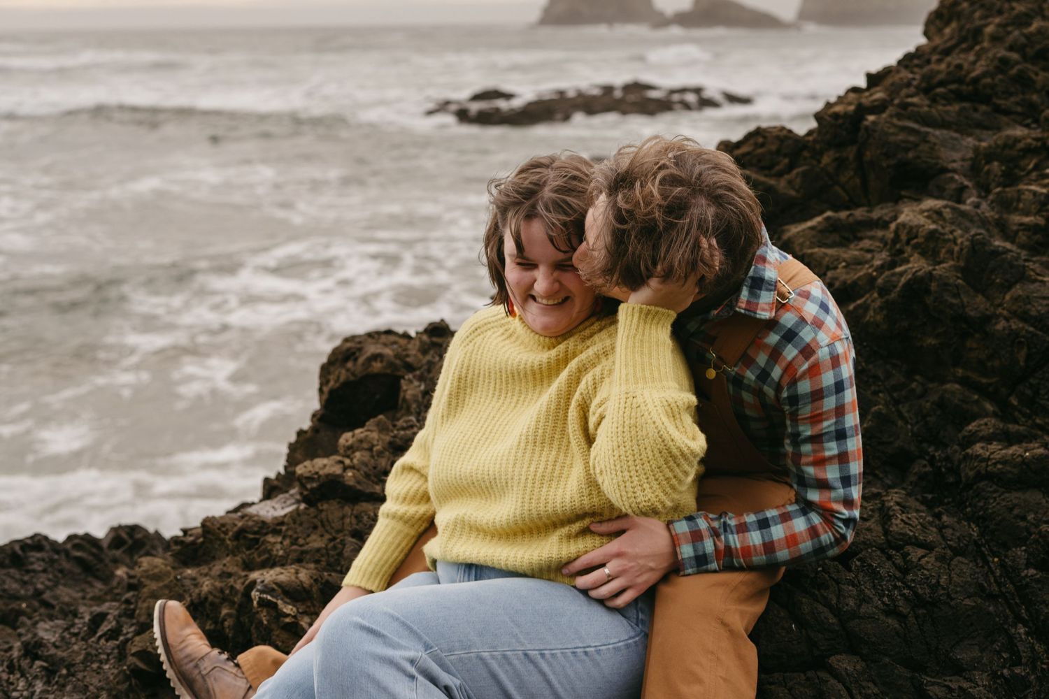 Two people share a romantic moment on rugged coastal rocks with crashing waves in background.