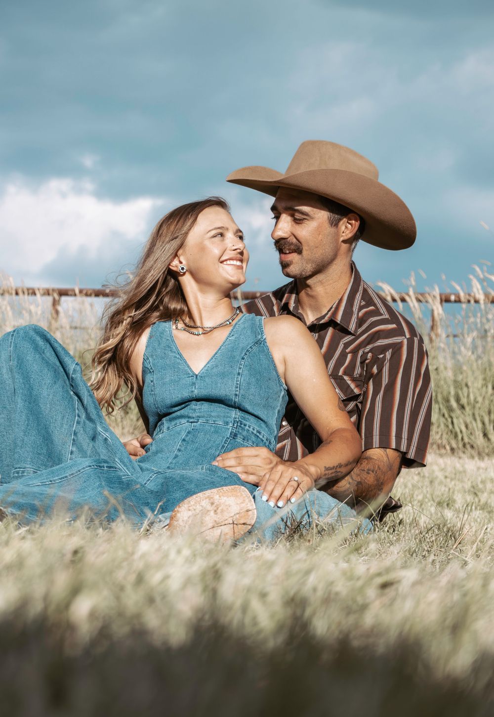 A couple in western attire share romantic moments while laying in tall grass under a blue sky.