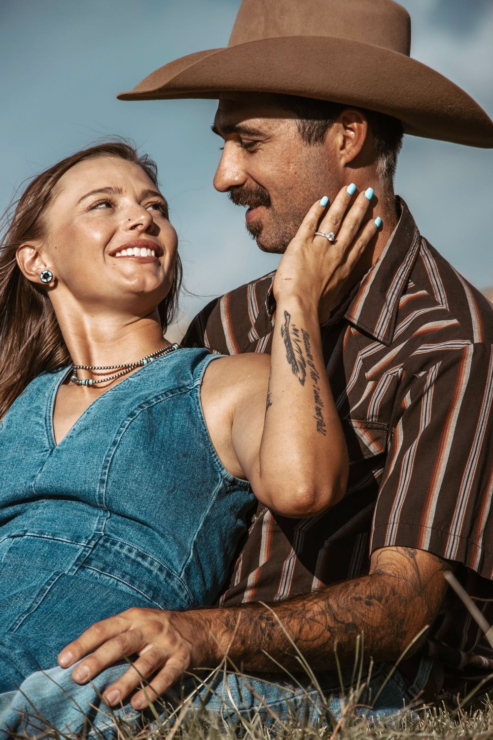 A couple in western attire sharing intimate moments while laying in grass during a sunny afternoon photoshoot.