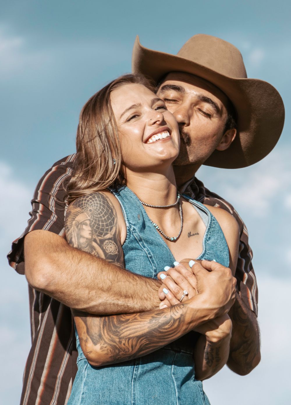 A couple shares a tender moment outdoors, with one wearing a cowboy hat and the other in a denim top against a blue sky.
