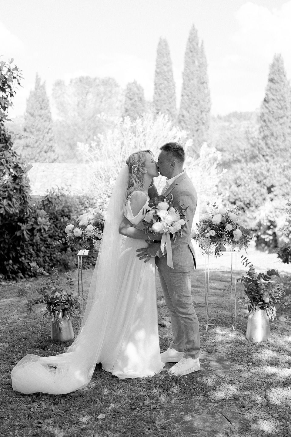 bride and groom kissing. They're standing between flower decorations at their Tuscan outdoor wedding during the ceremony