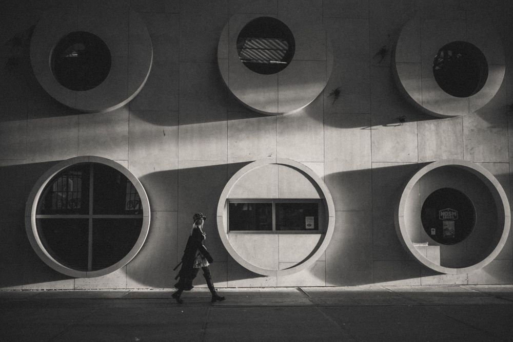 Person walking through a minimalist concrete hallway with circular windows creating dramatic shadows.