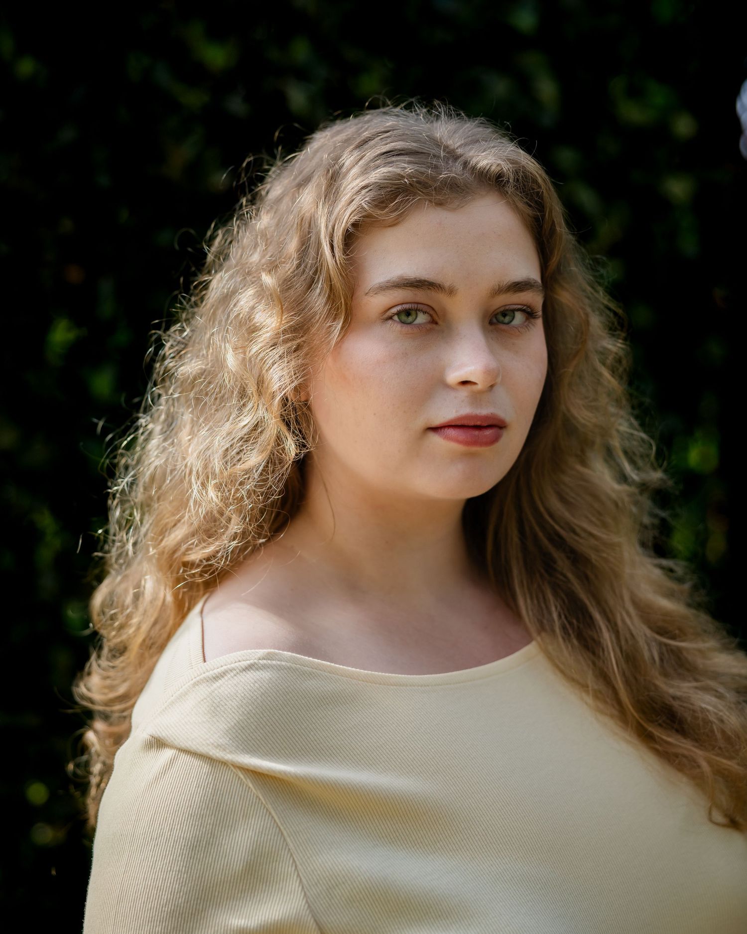 Young woman with curly blonde hair and green eyes wearing a yellow top, posing outdoors near green foliage.