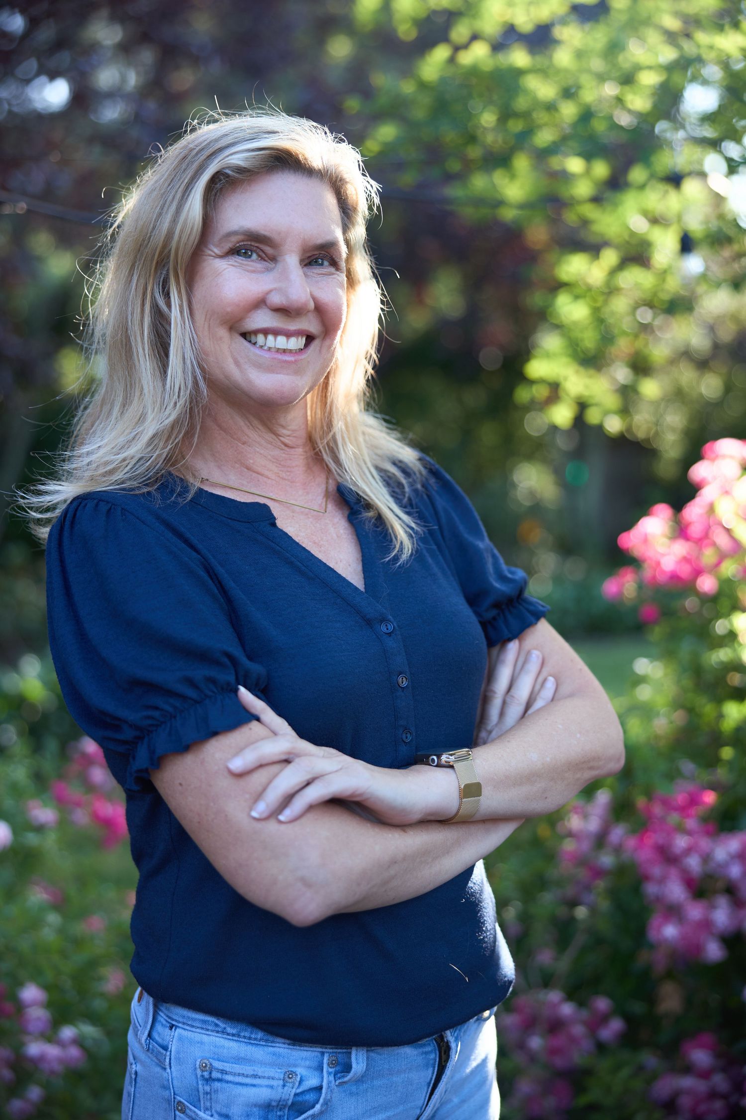 Woman in navy blue top poses confidently with arms crossed in garden with blooming pink flowers in background.