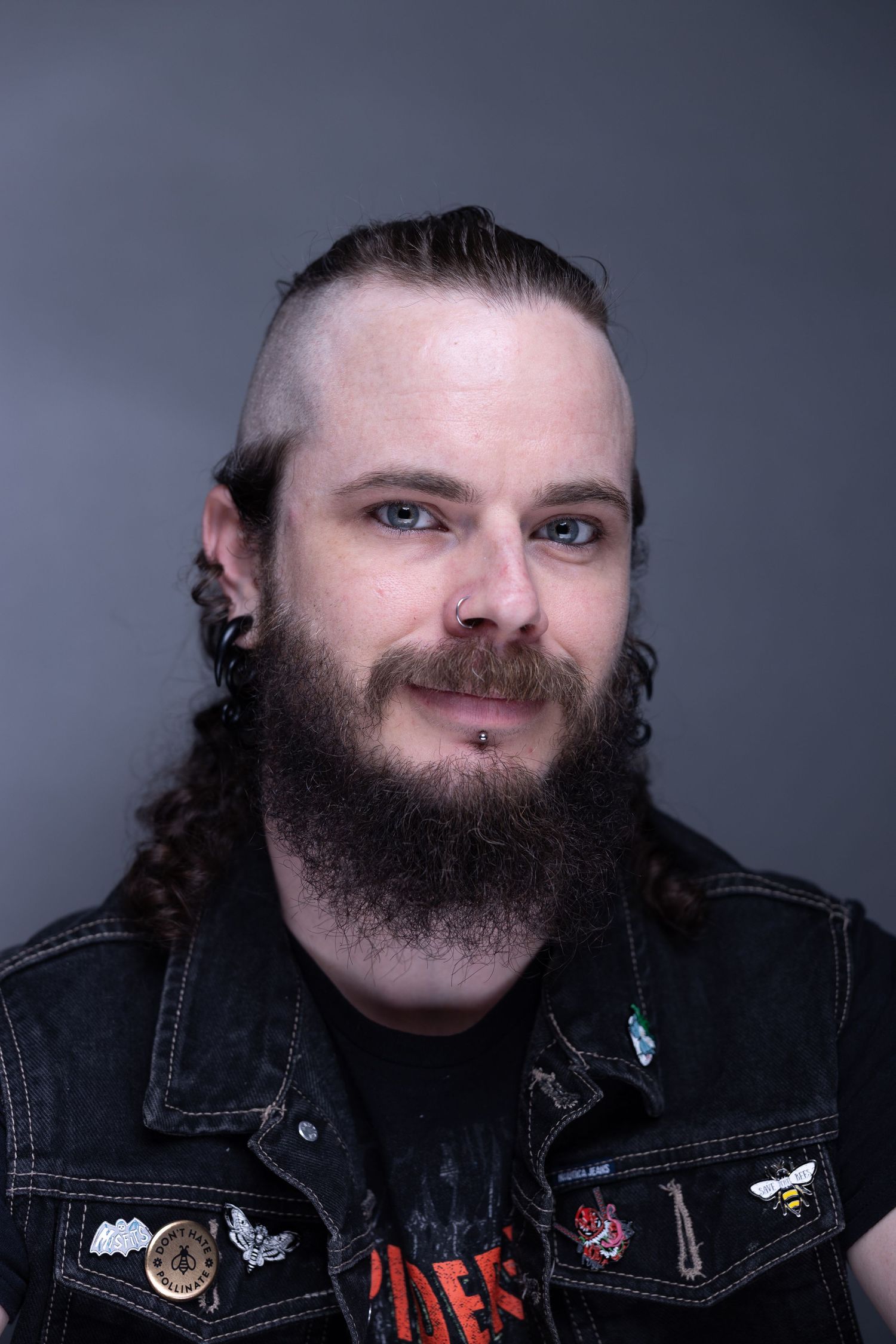 A bearded person wearing a denim vest with patches smiles against a dark studio backdrop.