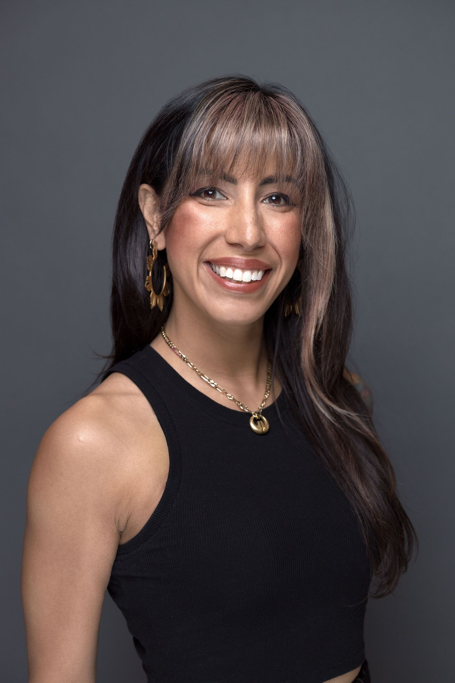 Professional portrait of someone wearing a black sleeveless top with long dark hair and bangs, smiling against a gray background.