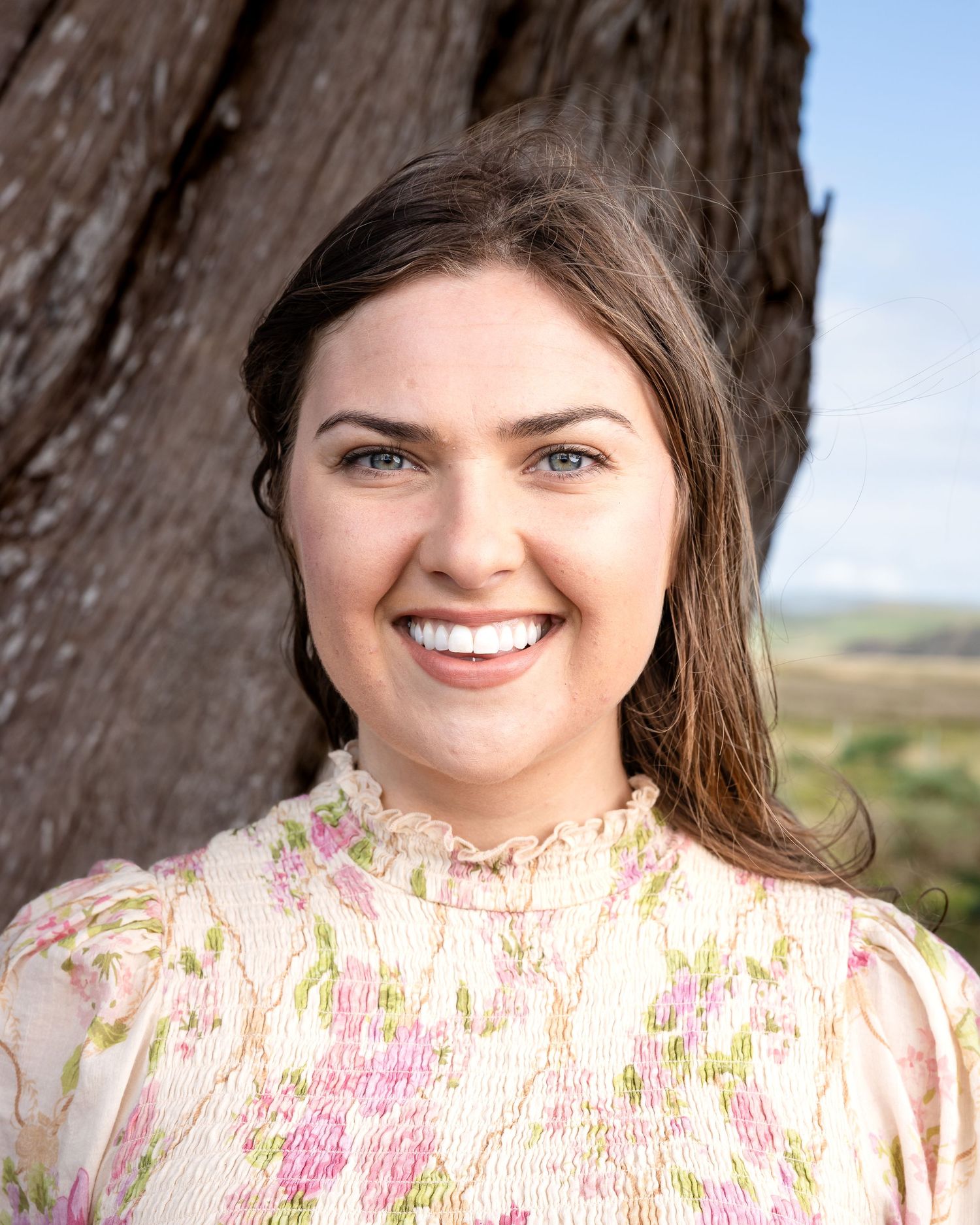 Smiling person wearing floral pink blouse poses near rustic tree trunk in outdoor setting with natural landscape behind them.