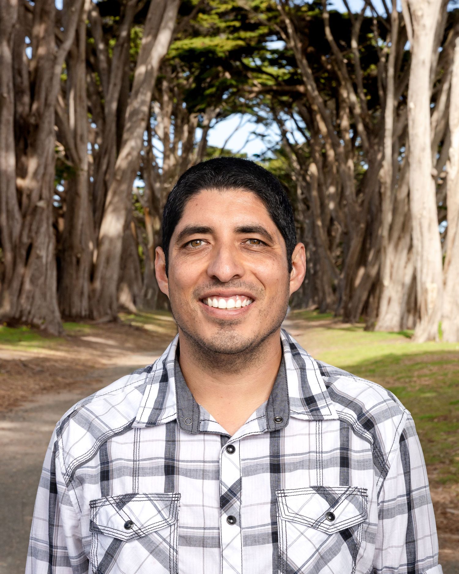 A smiling person in a plaid shirt stands on a tree-lined path with tall cypress trees forming a natural tunnel.