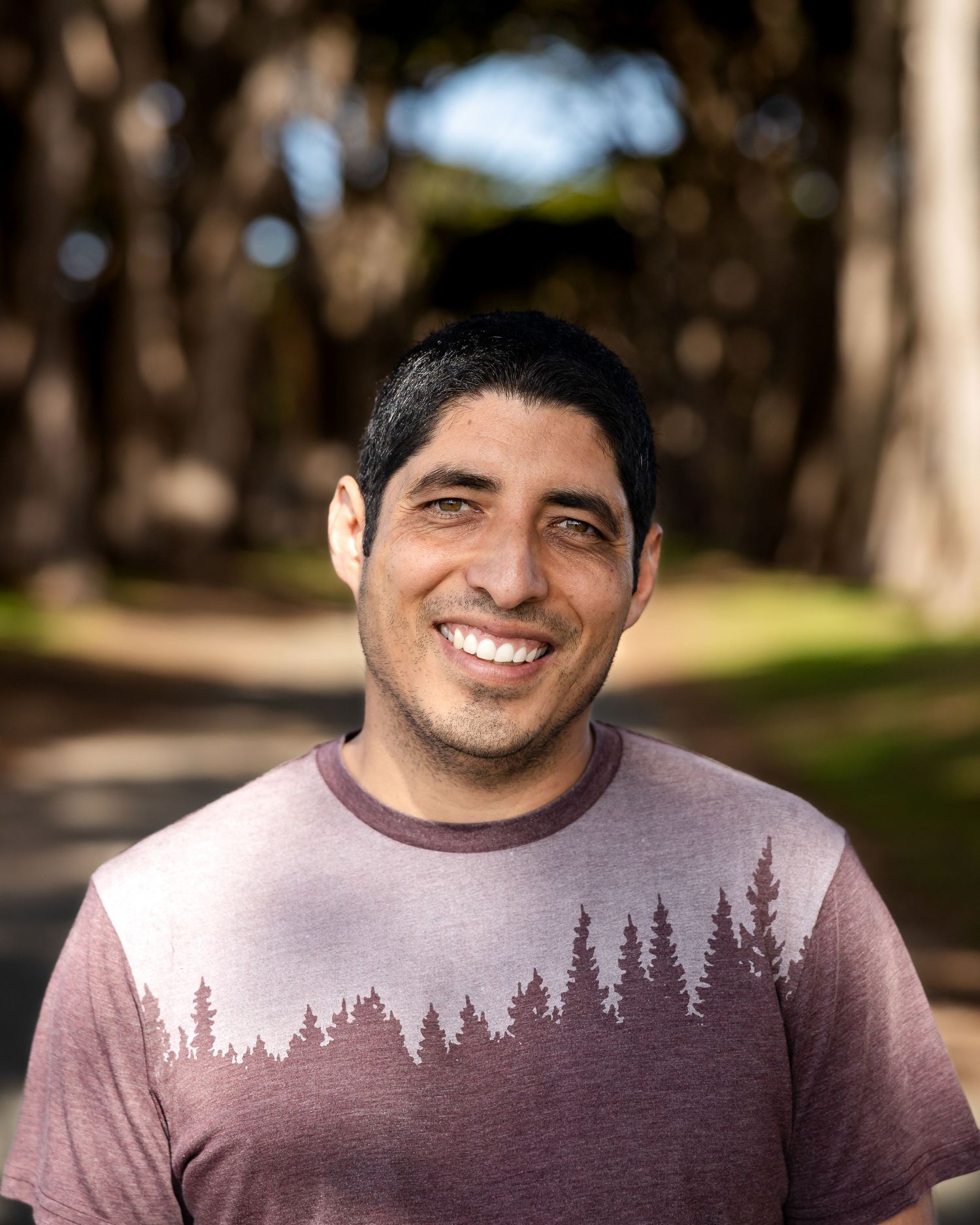 Smiling person wearing a mauve t-shirt with forest silhouette design outdoors in front of blurred trees.