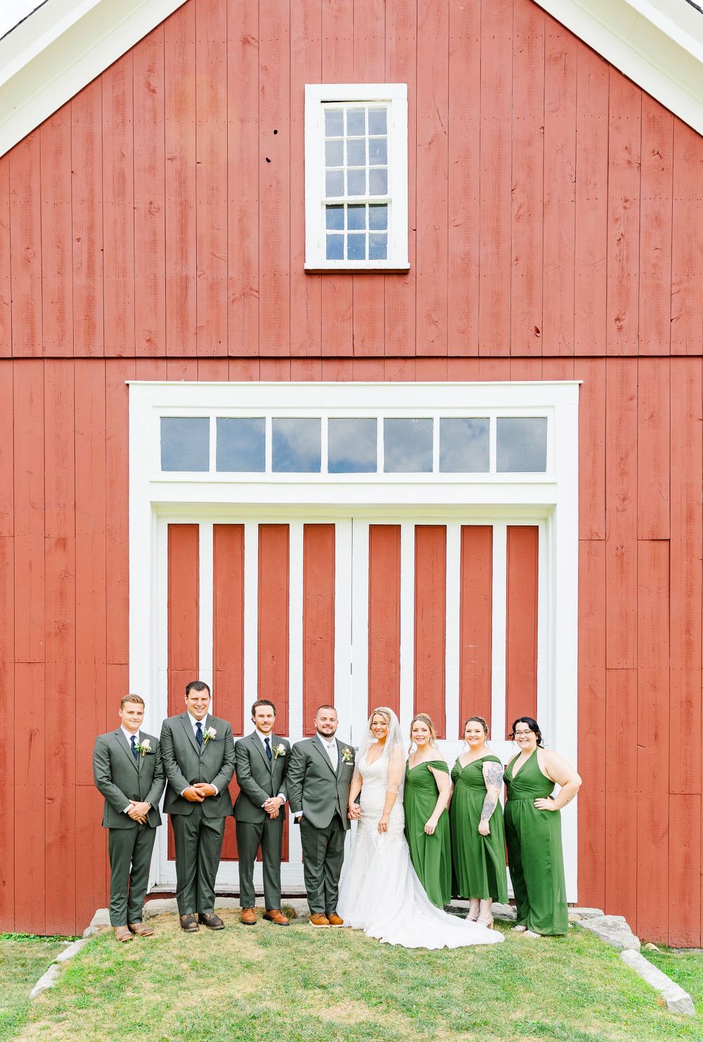 Wedding party in green and gray attire lined up against rustic red barn with white trim windows.