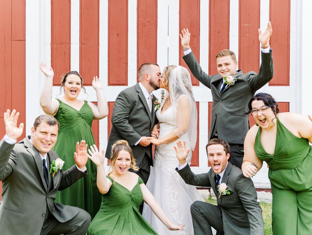 Wedding party in emerald green dresses and gray suits poses joyfully against red barn exterior.
