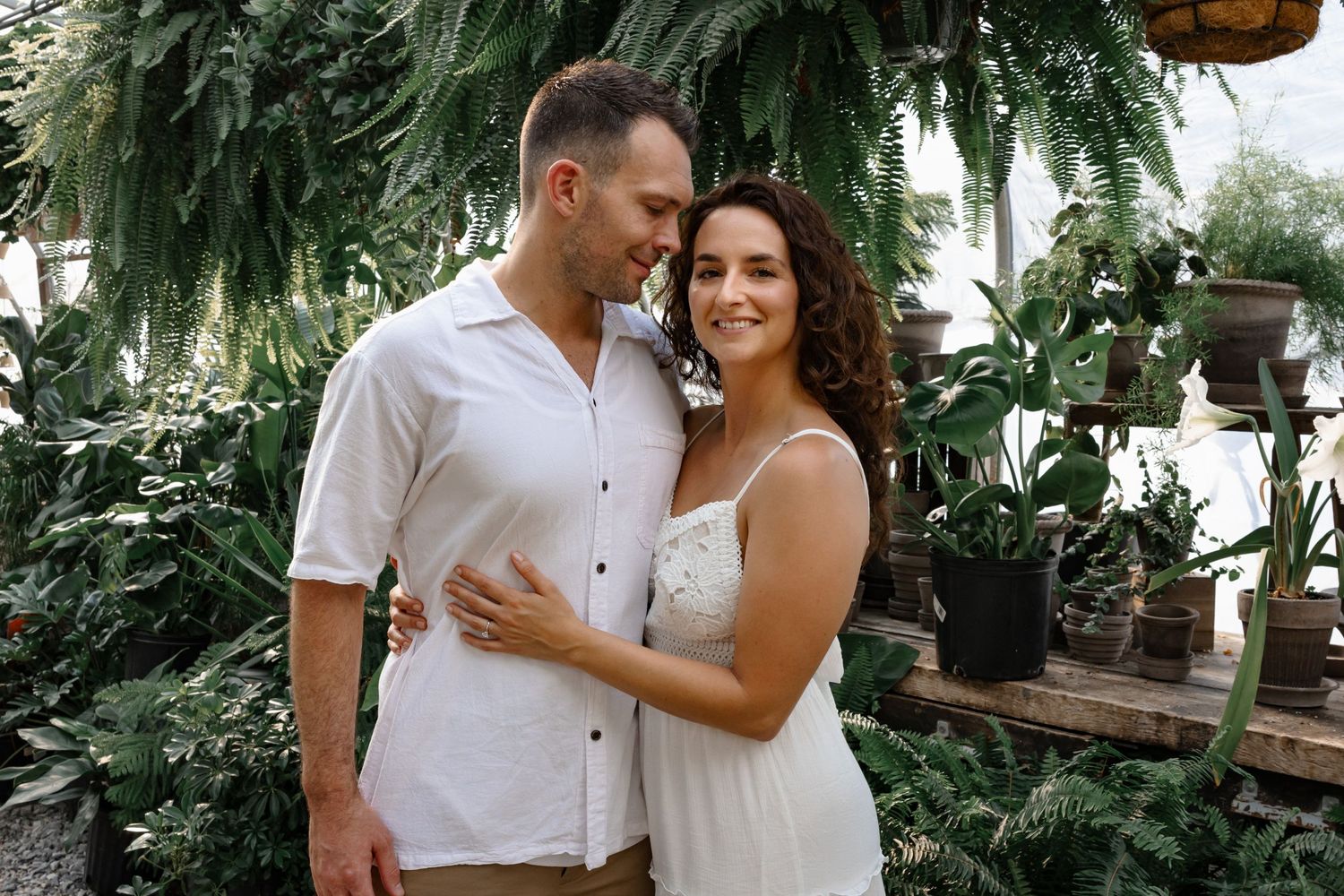Romantic greenhouse engagement photo with lush ferns and plants in background.