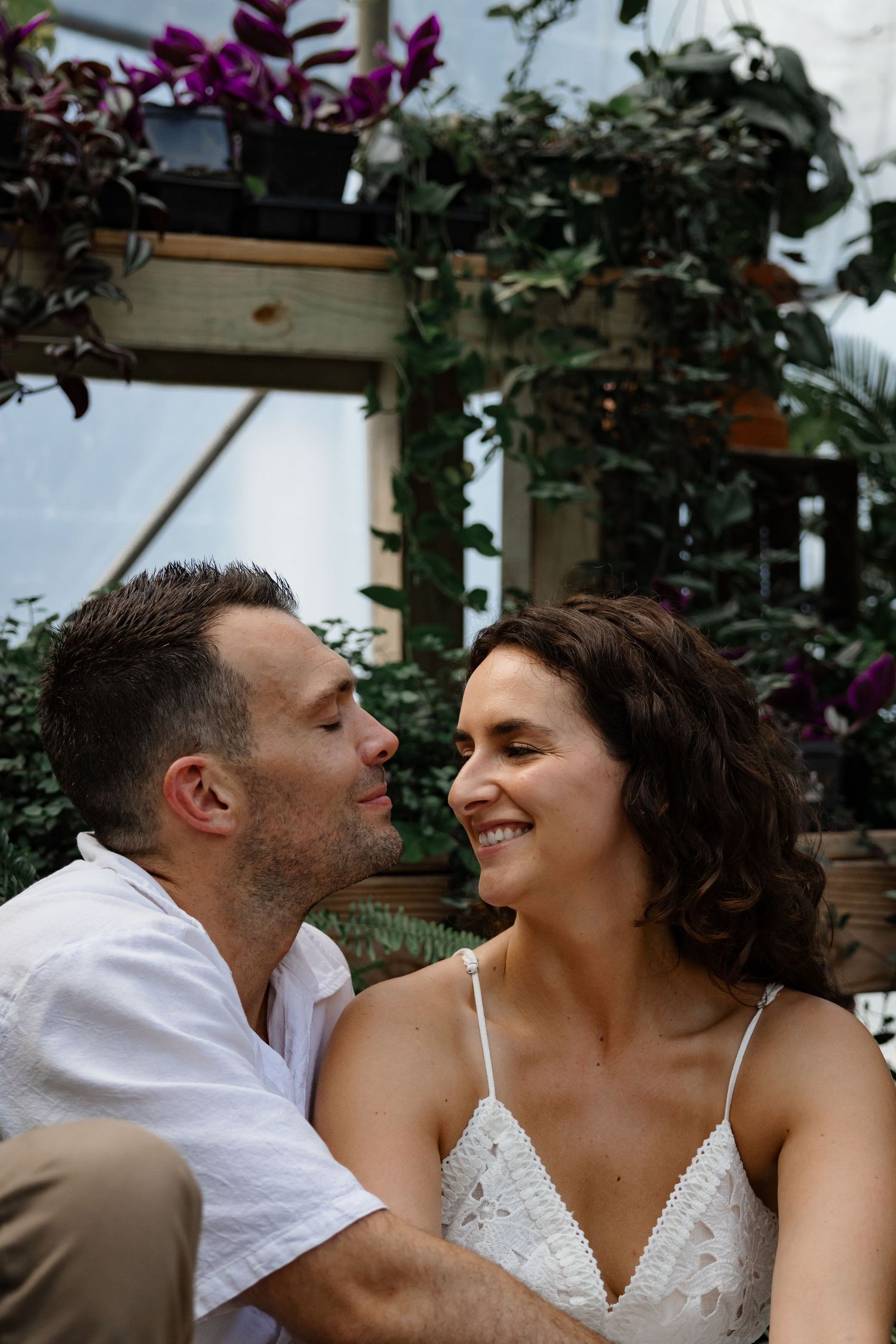 Tender moment shared between couple surrounded by greenhouse plants and purple flowers.