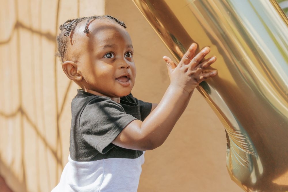 A child reaches up towards a gold balloon against a beige wall with sunlight casting geometric shadows.