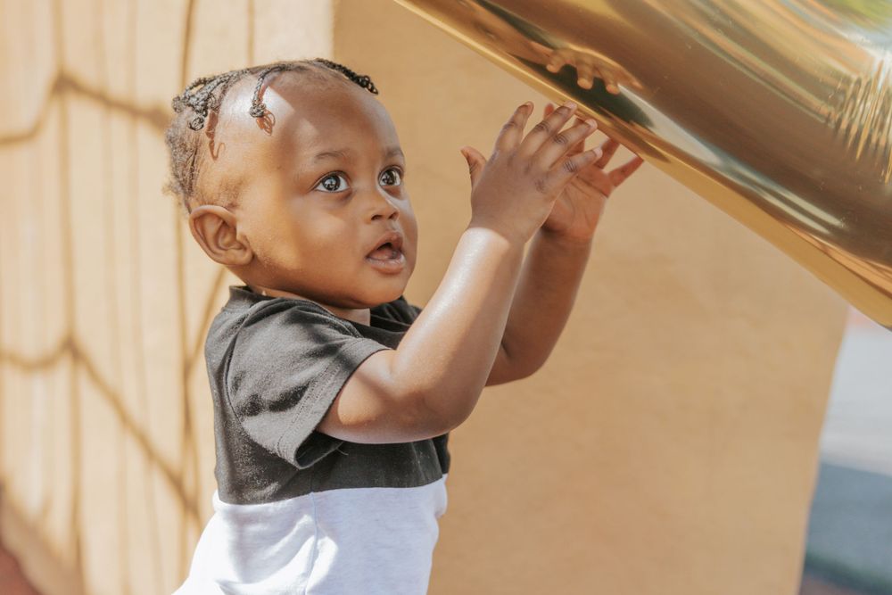 A small child reaches up towards a shelf while wearing a gray shirt and white garment.