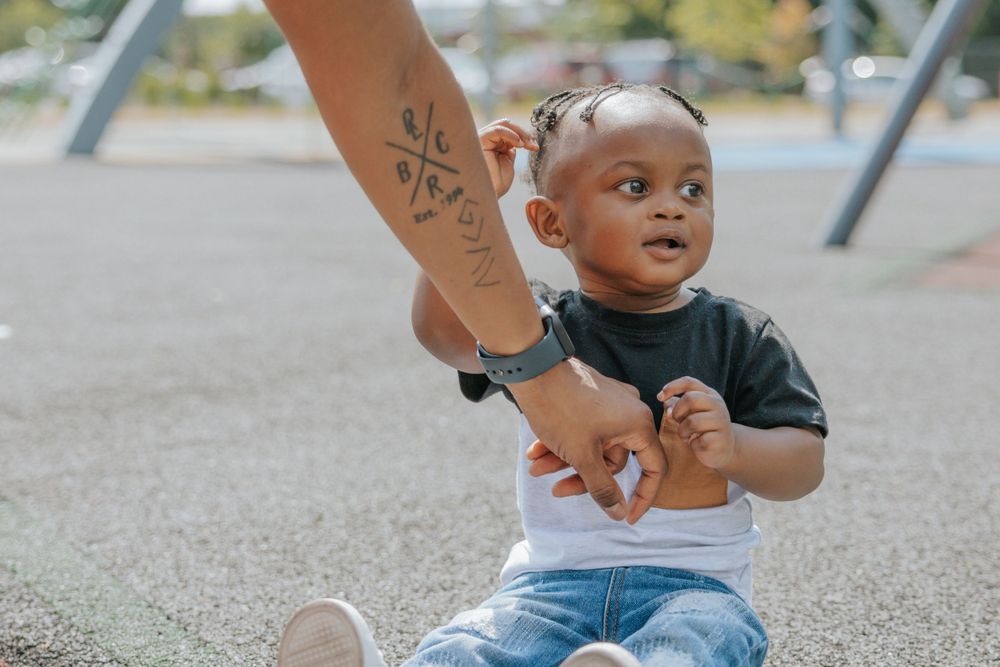 A toddler on a playground reaches up while being supported by an adult arm with a tattoo.