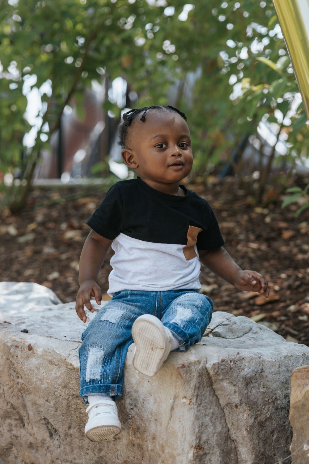 A child in a two-tone shirt and jeans sits on a large rock in a garden setting.